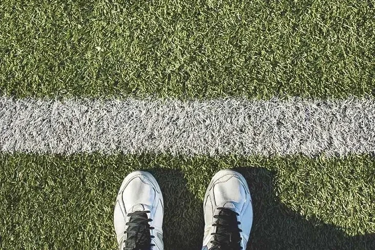Overhead view of white sneakers standing behind a white painted line on green artificial turf sports field.