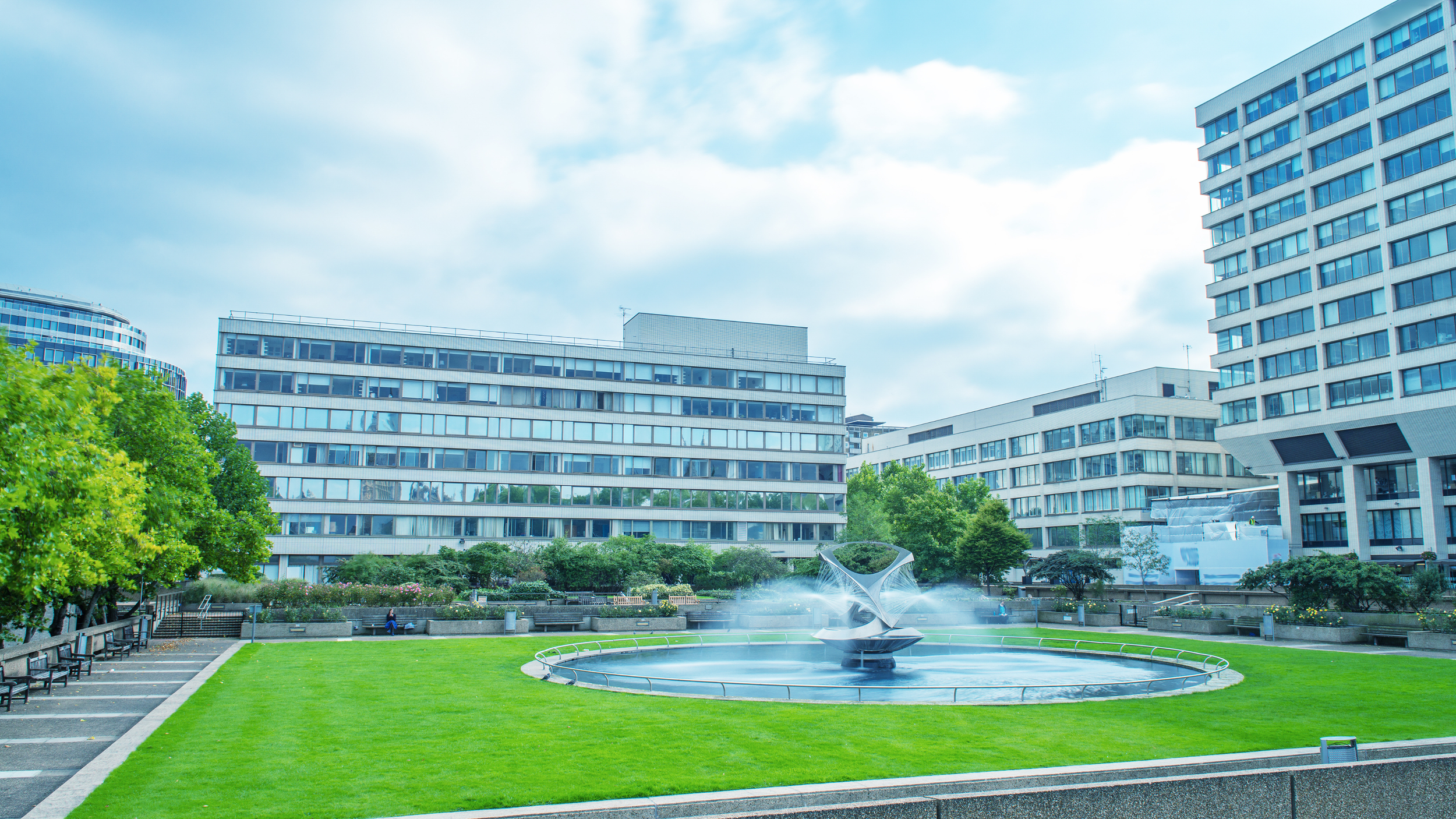 A modern metal fountain sprays water in a circular pool on a green lawn, surrounded by large office buildings.