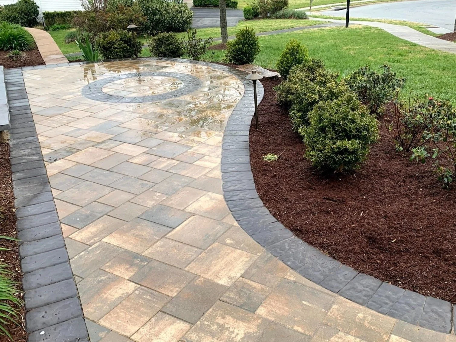 Elegant paver patio featuring a circular stone inlay and dark border, next to a freshly mulched garden bed.