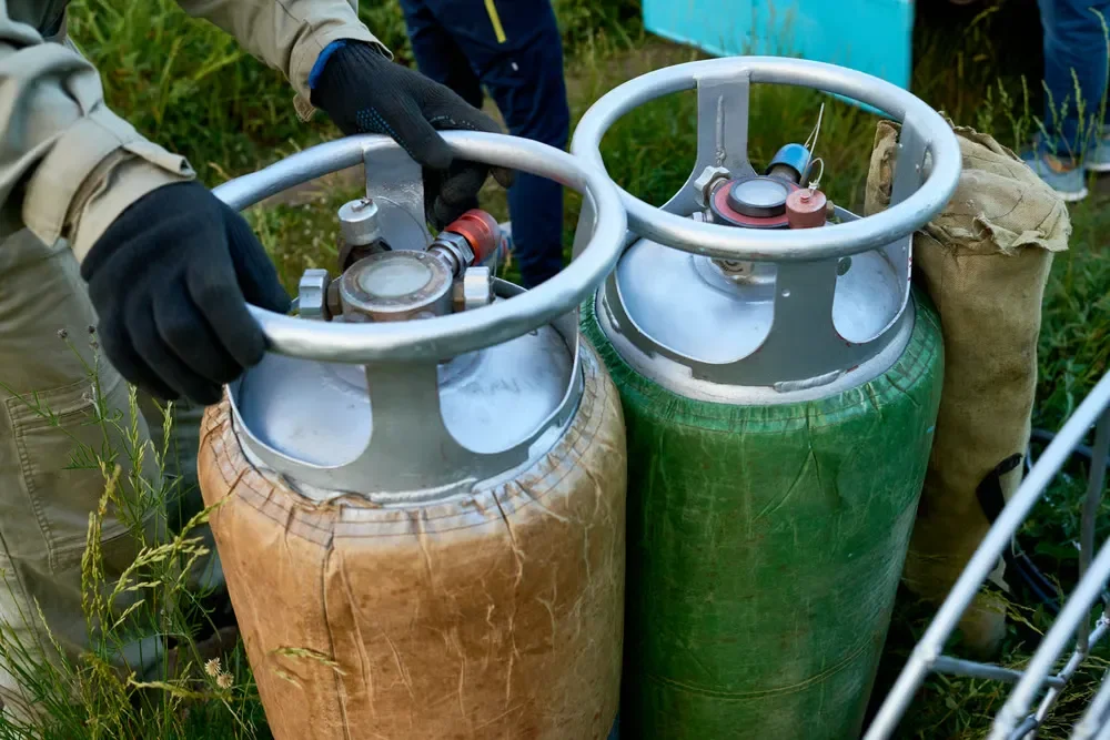 Gloved hands adjusting the top valve of a lightweight brown composite propane tank standing next to a green gas cylinder.