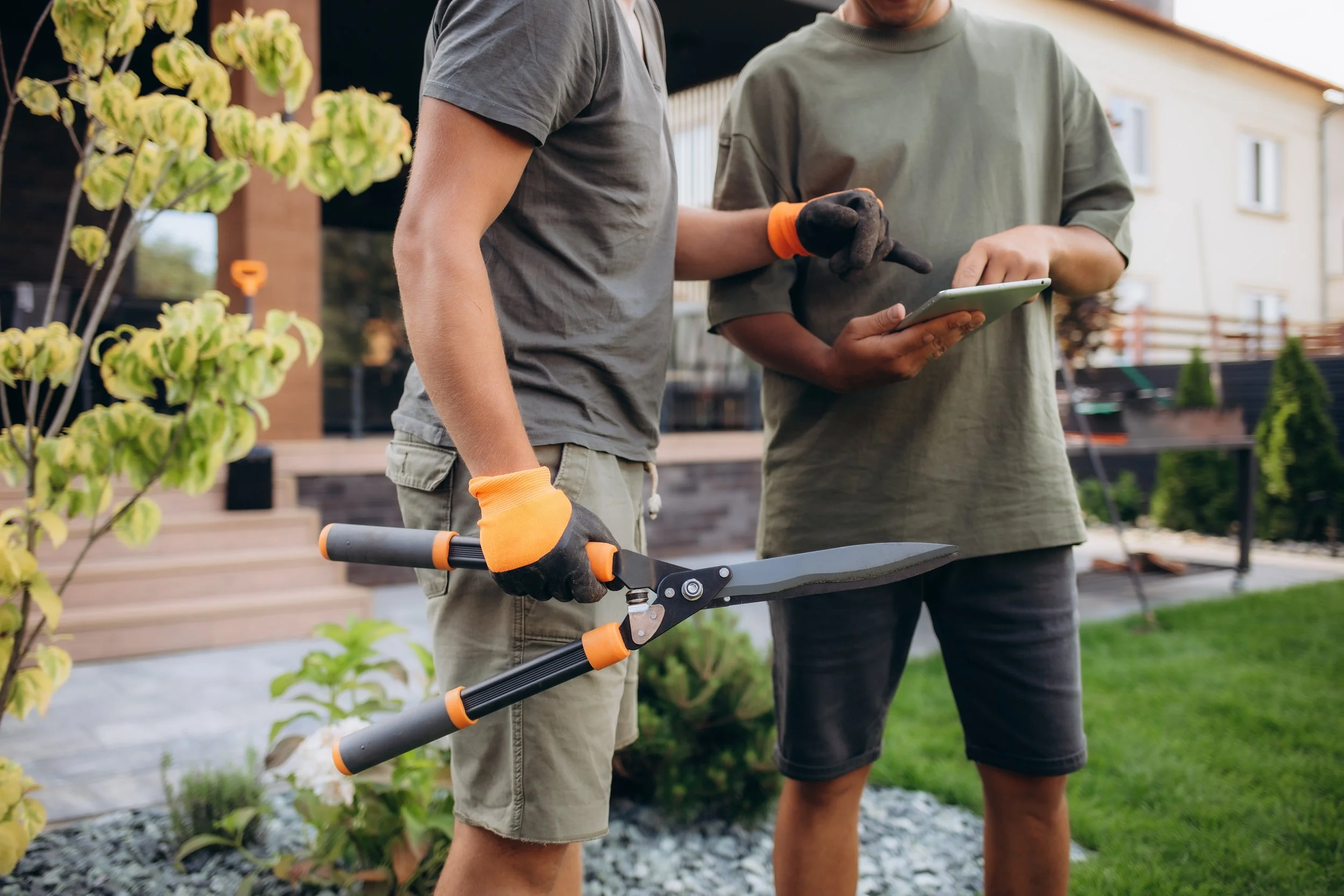 A midsection view of two landscapers, one holding large shears and the other holding a tablet.