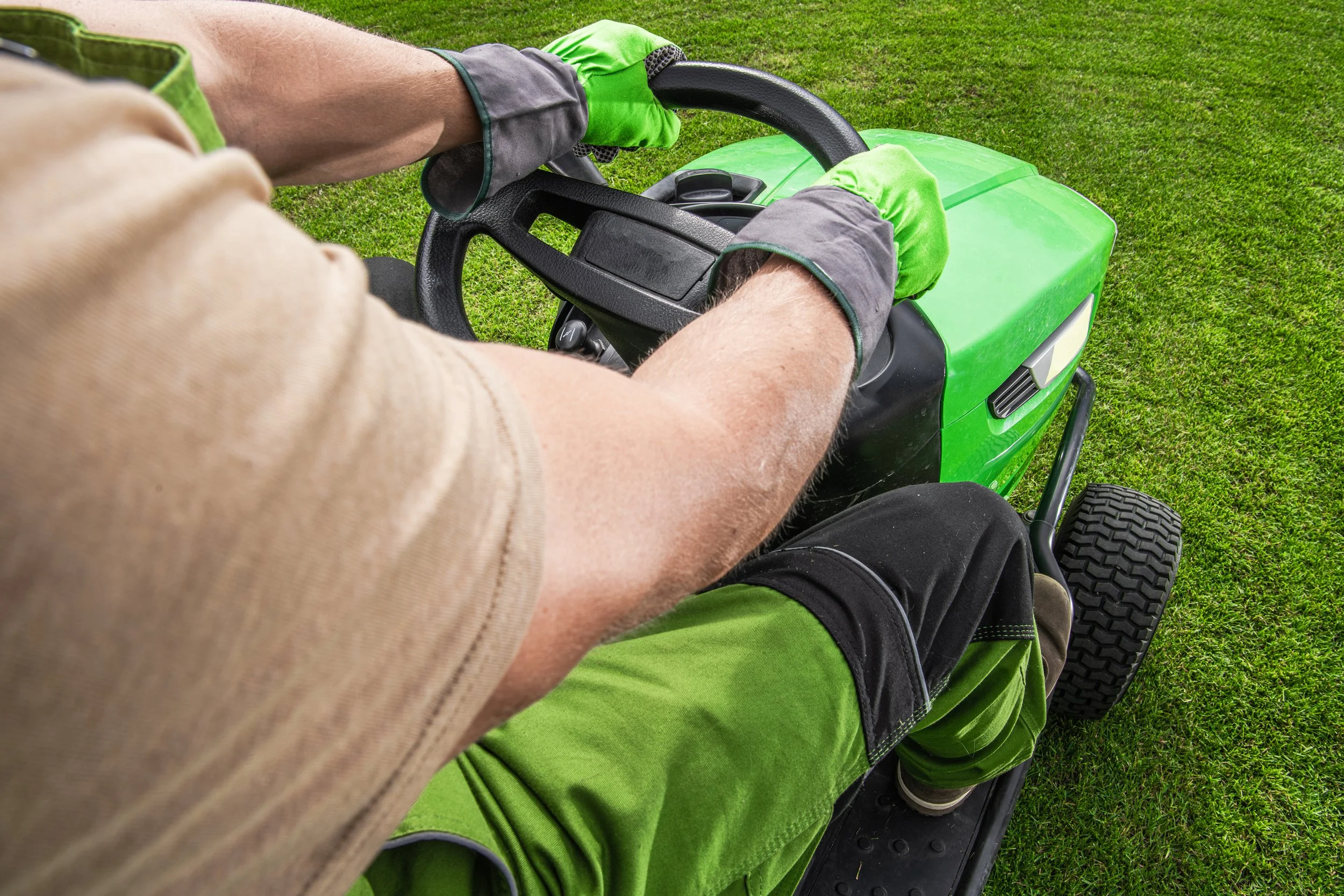 A person in green gloves and workwear operates a bright green riding lawn mower across a lush, grassy field.