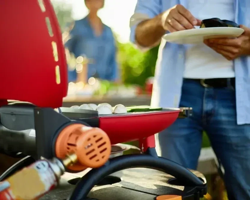 Close-up of a red portable gas grill cooking food at a backyard barbecue with a man holding a plate.