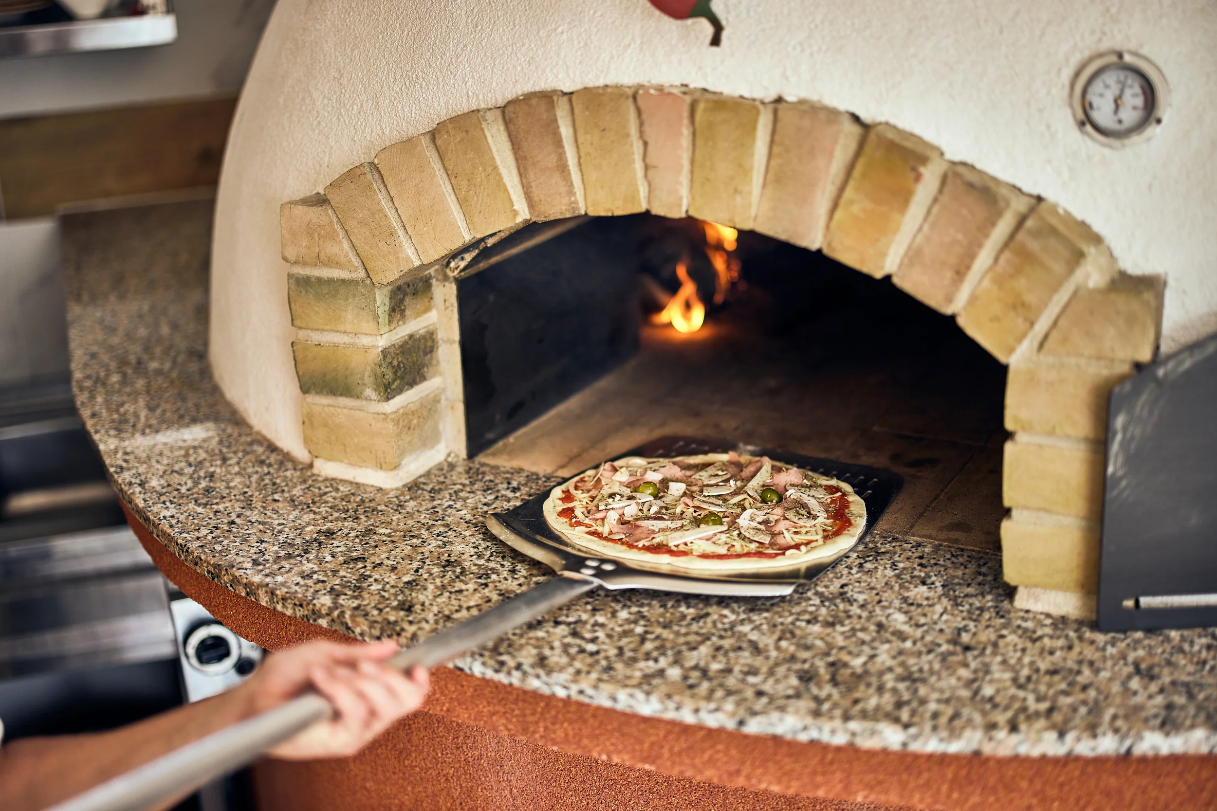 Chef sliding a pizza on a metal peel into a wood-fired brick pizza oven with a granite counter