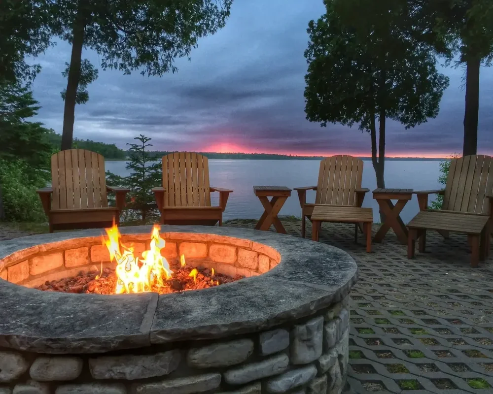 Outdoor circular stone fire pit burning on a patio with wooden Adirondack chairs overlooking a lake at dusk.