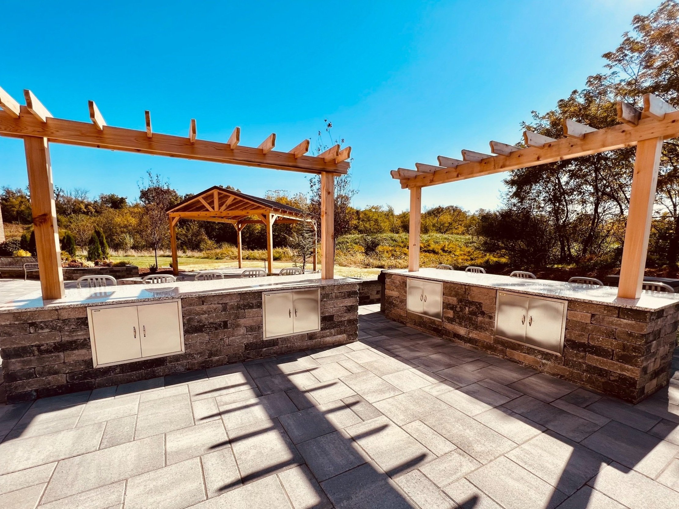 Custom outdoor kitchen with stone cabinets and wooden pergolas on a modern paver patio overlooking a lawn.
