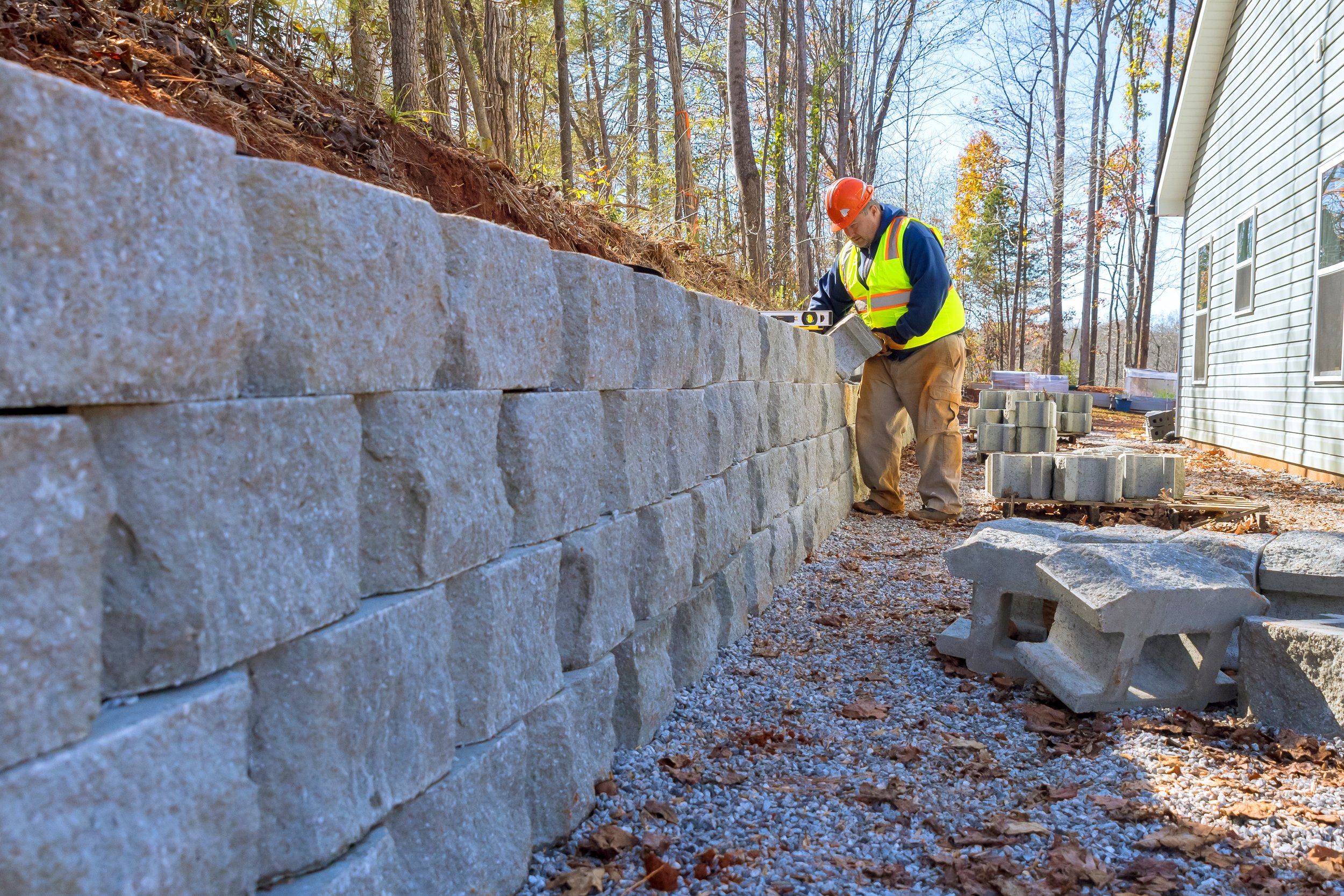 A contractor in a hard hat and safety vest installing stone blocks for a large backyard retaining wall.