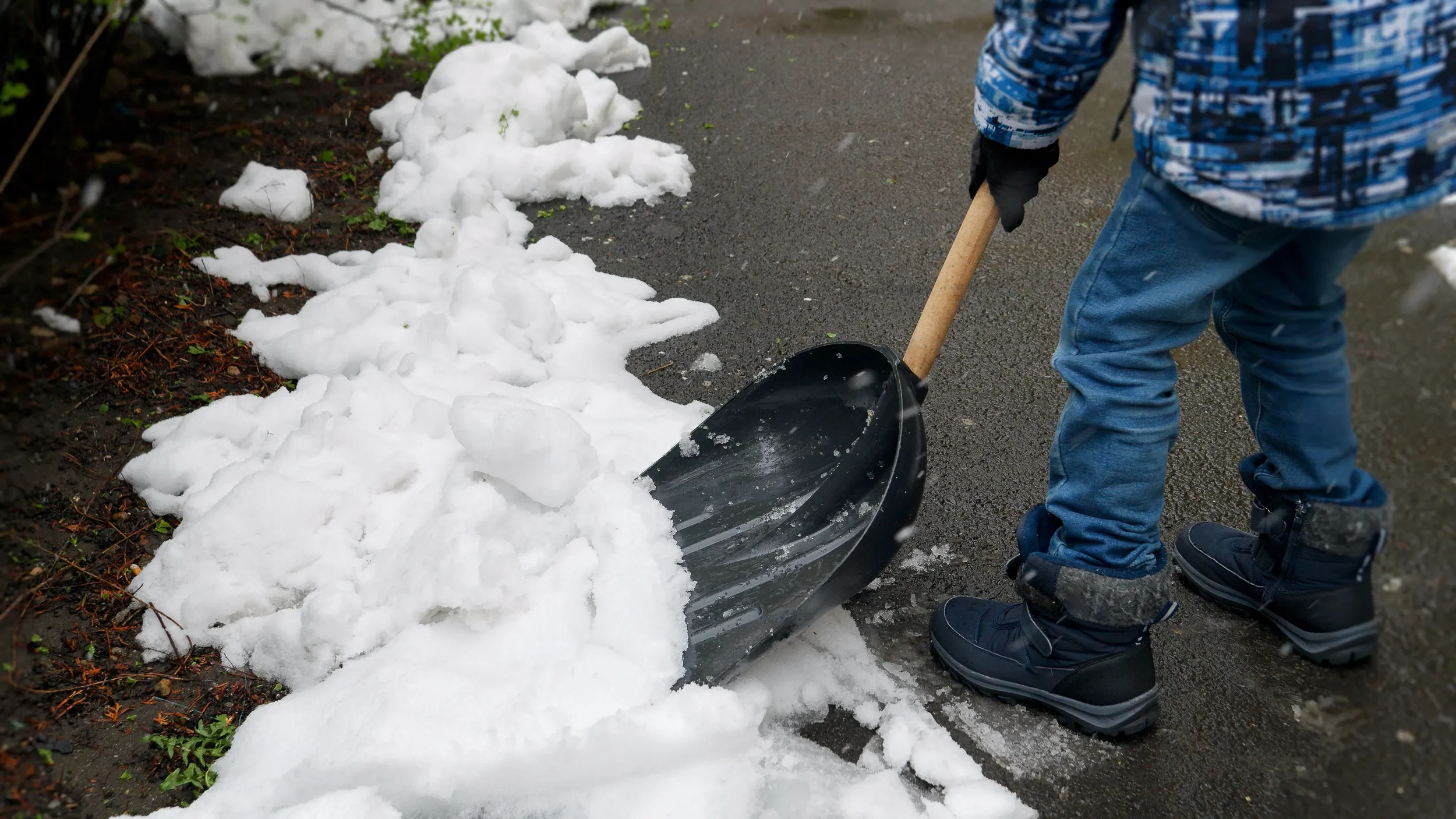 Close-up of a person using a black shovel to clear a path through thick white snow on a paved surface.
