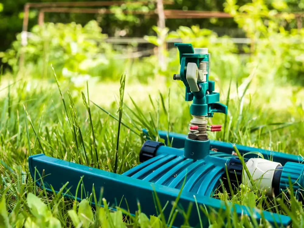 Close-up of a green plastic portable lawn sprinkler sitting on lush green grass in a backyard.