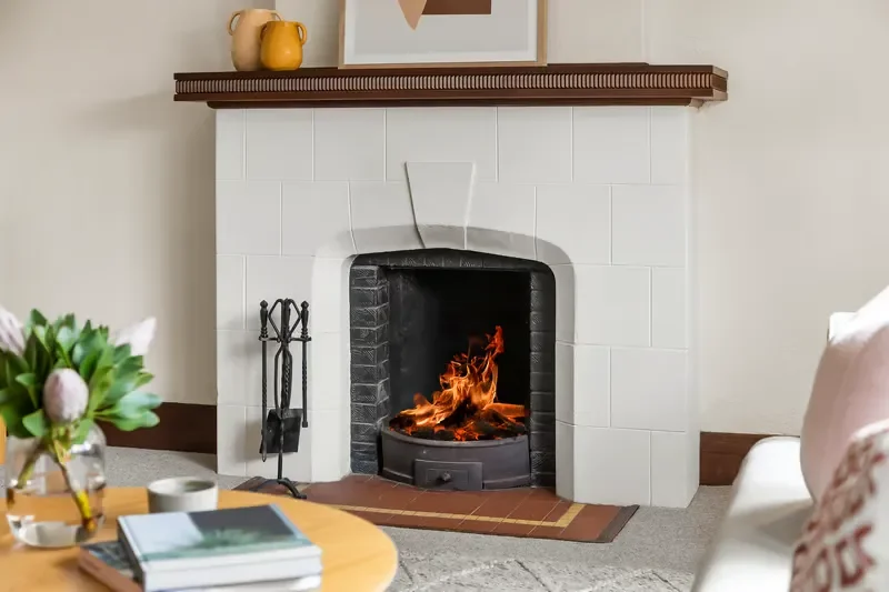 Burning wood fire in a white tiled fireplace with a dark wood mantel and black iron tools in a cozy living room.