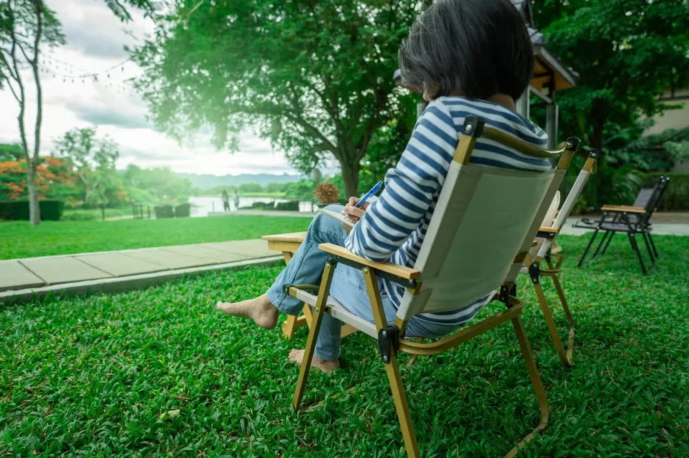 A person in a striped shirt sits on a folding lawn chair writing on a notepad in a green backyard grass lawn.