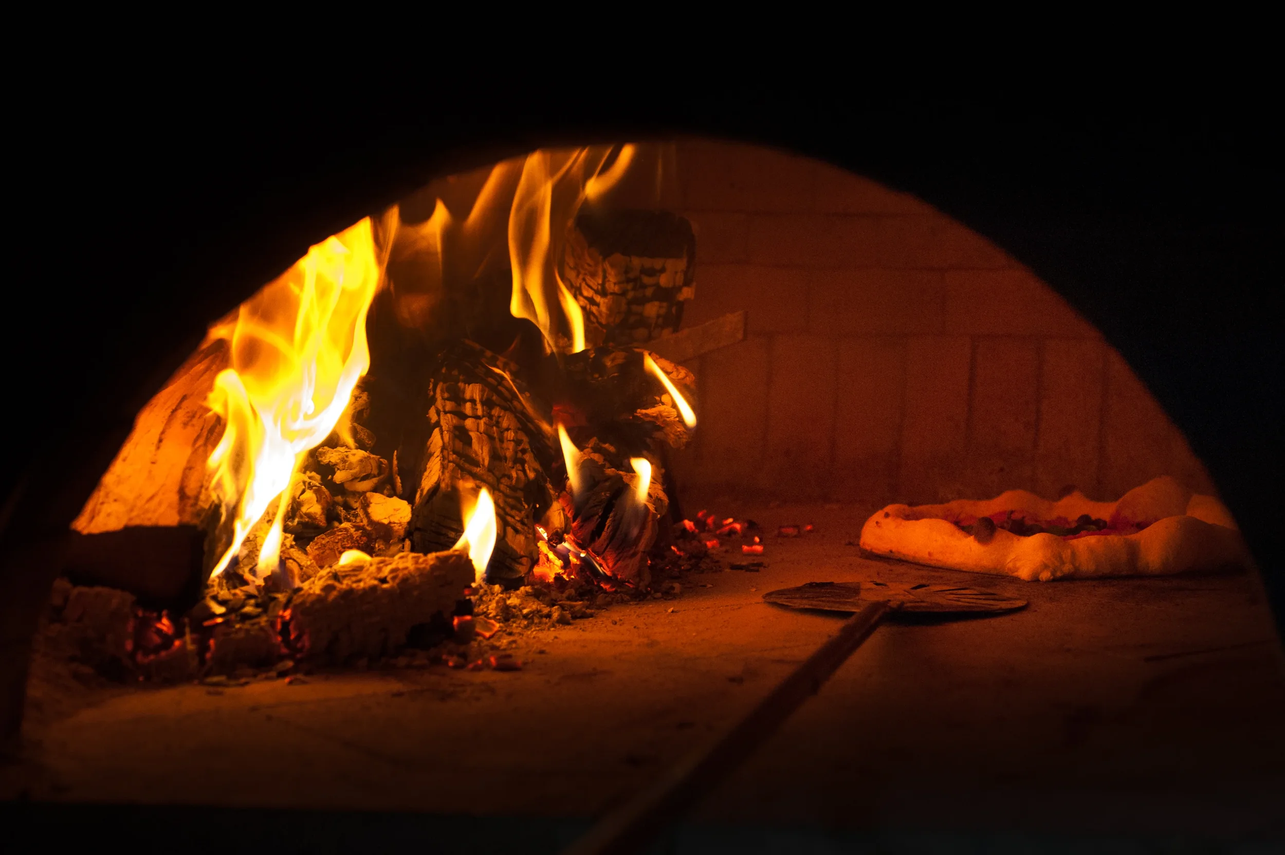 Pizza baking beside a blazing wood fire and glowing embers inside a dark brick pizza oven