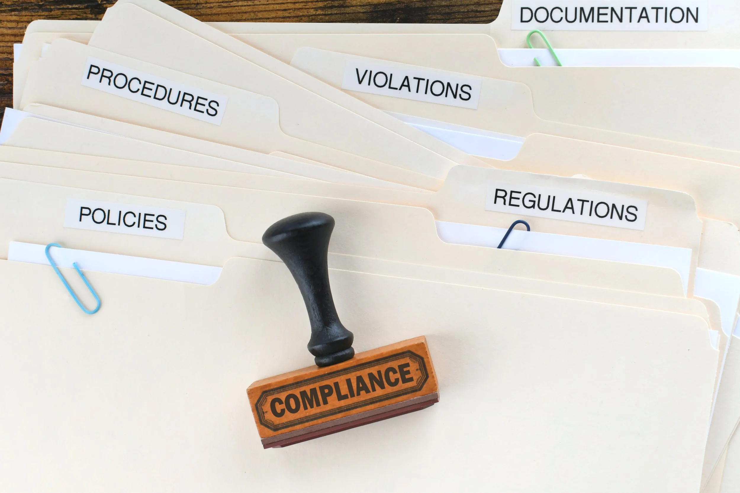 Manila folders labeled Policies, Procedures, Violations, Regulations, and Documentation with a "Compliance" stamp in the foreground.