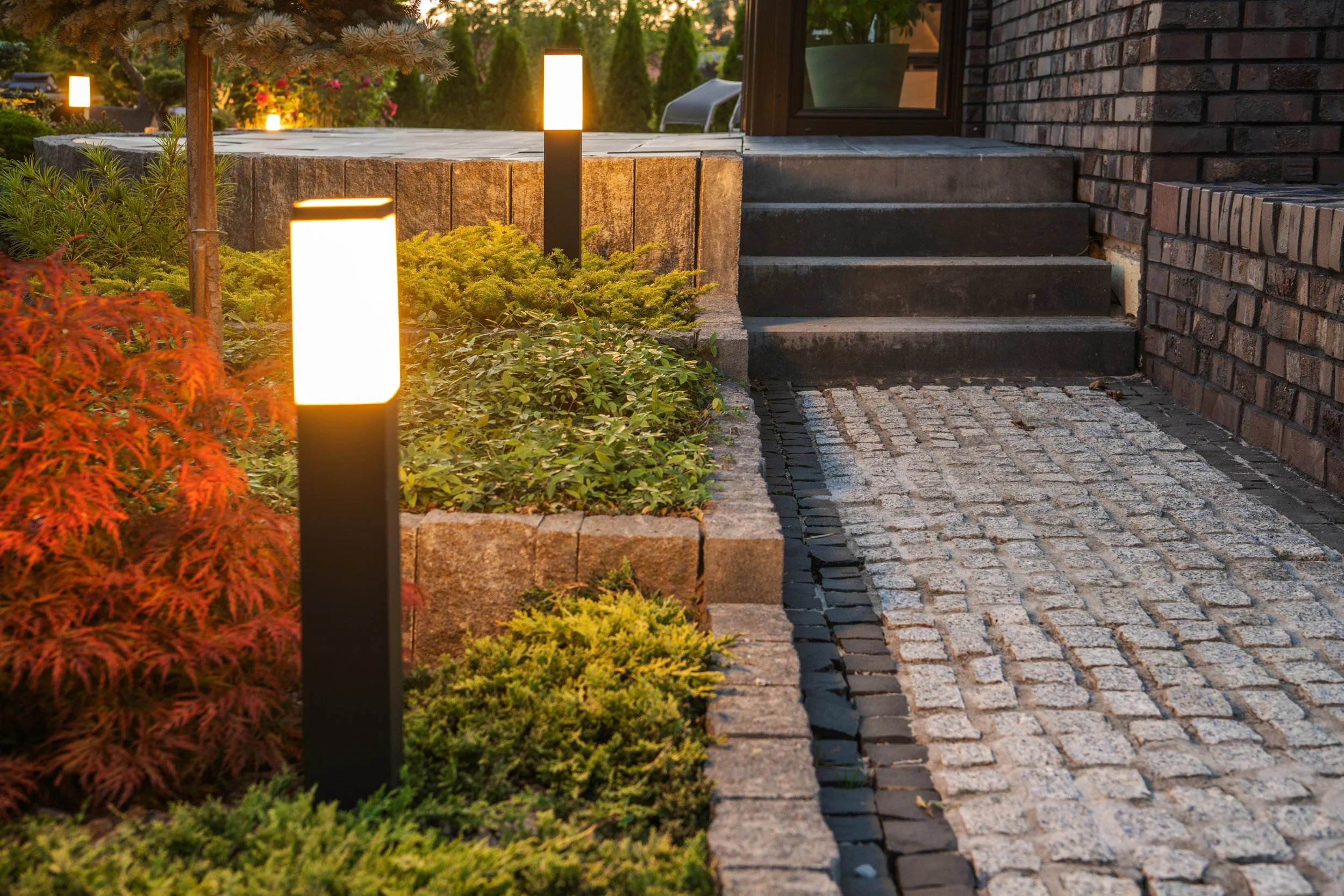 Modern black bollard lights illuminating a stone walkway and garden beds near a brick house entrance.
