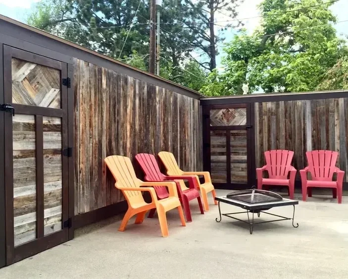 Colorful Adirondack chairs and a metal fire pit arranged on a concrete patio enclosed by rustic wooden fencing.
