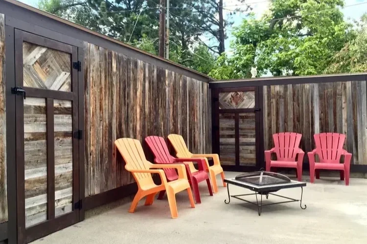 Rustic reclaimed wood privacy fence with a gate around a patio with a fire pit and colorful Adirondack chairs.