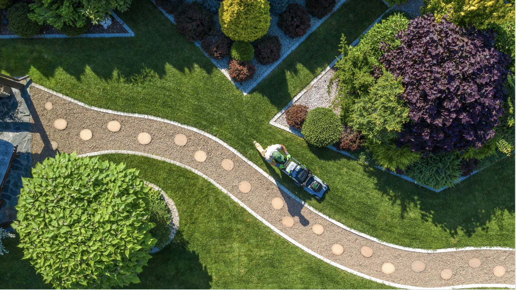 An aerial view shows a person pushing a lawn mower across a bright green lawn next to a winding gravel path with stepping stone