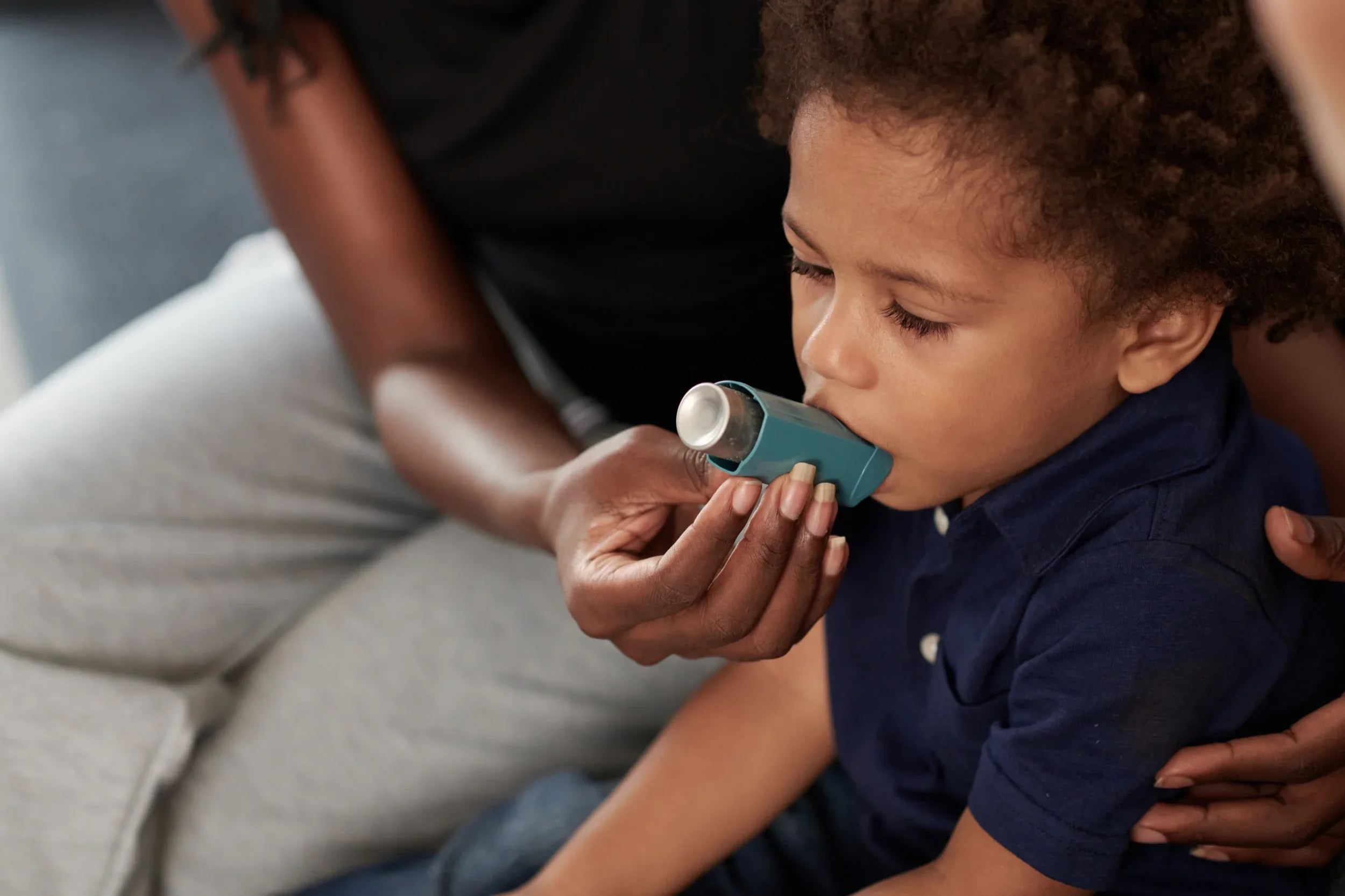 A young child receiving help from an adult to use a blue asthma inhaler for respiratory relief.