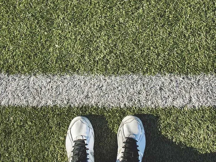 White athletic sneakers standing in front of a white boundary line on green artificial turf field grass.
