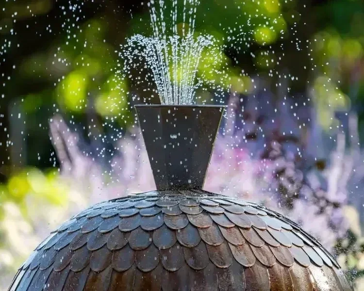 Decorative tiered dome garden water fountain spraying water droplets in a lush outdoor backyard setting.