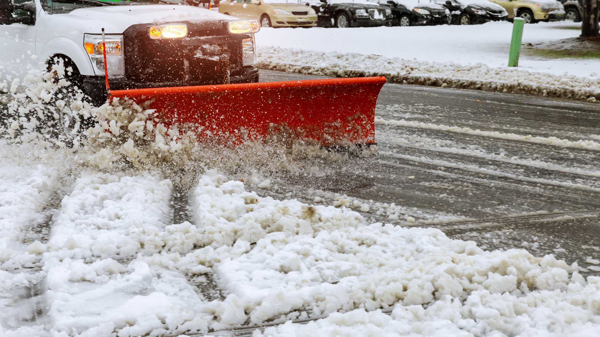 A white pickup truck with a large red snow plow attachment clearing heavy slush and snow from a commercial parking lot.