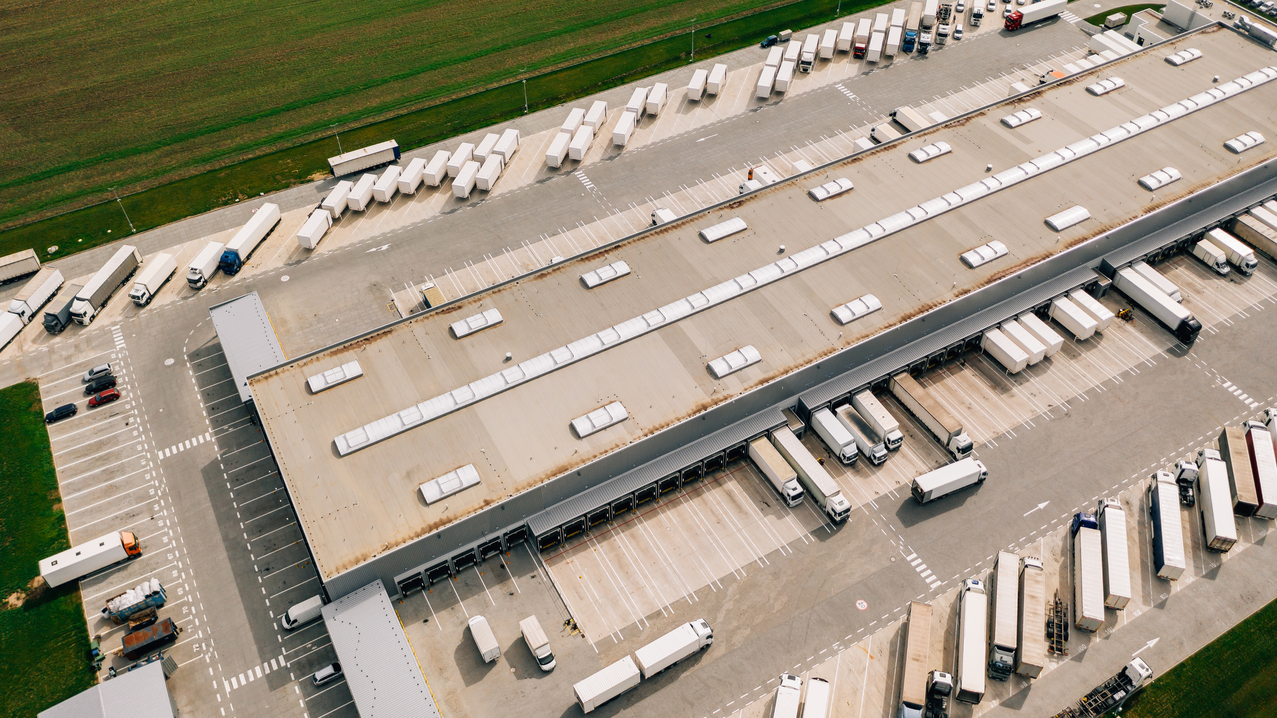 An aerial view of a large logistics warehouse with numerous white semi-trucks parked at loading docks and in lots.