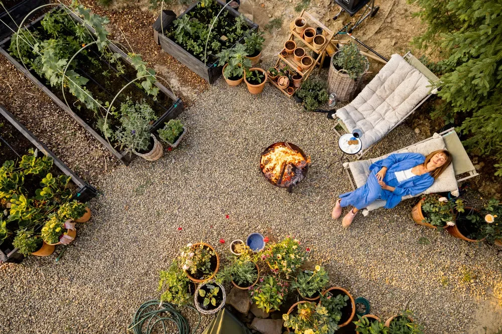Top-down view of a gravel backyard patio with a fire pit, lounge chairs, potted plants, and raised garden beds.