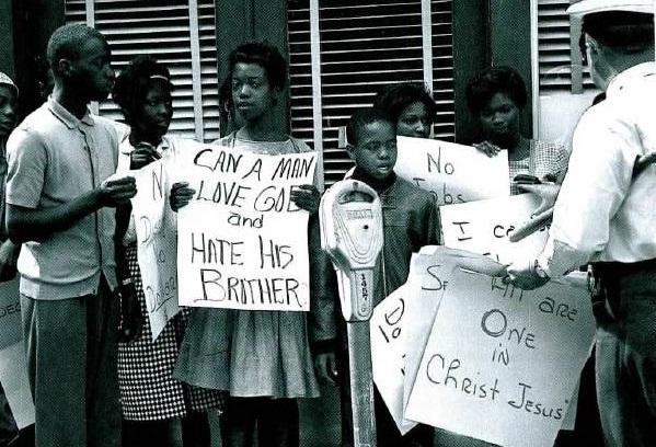 A young black woman holds a sign that reads: "Can a Man Love God and Hate his Brother?" A white police officer takes away a sign that reads: "All are One in Christ Jesus."