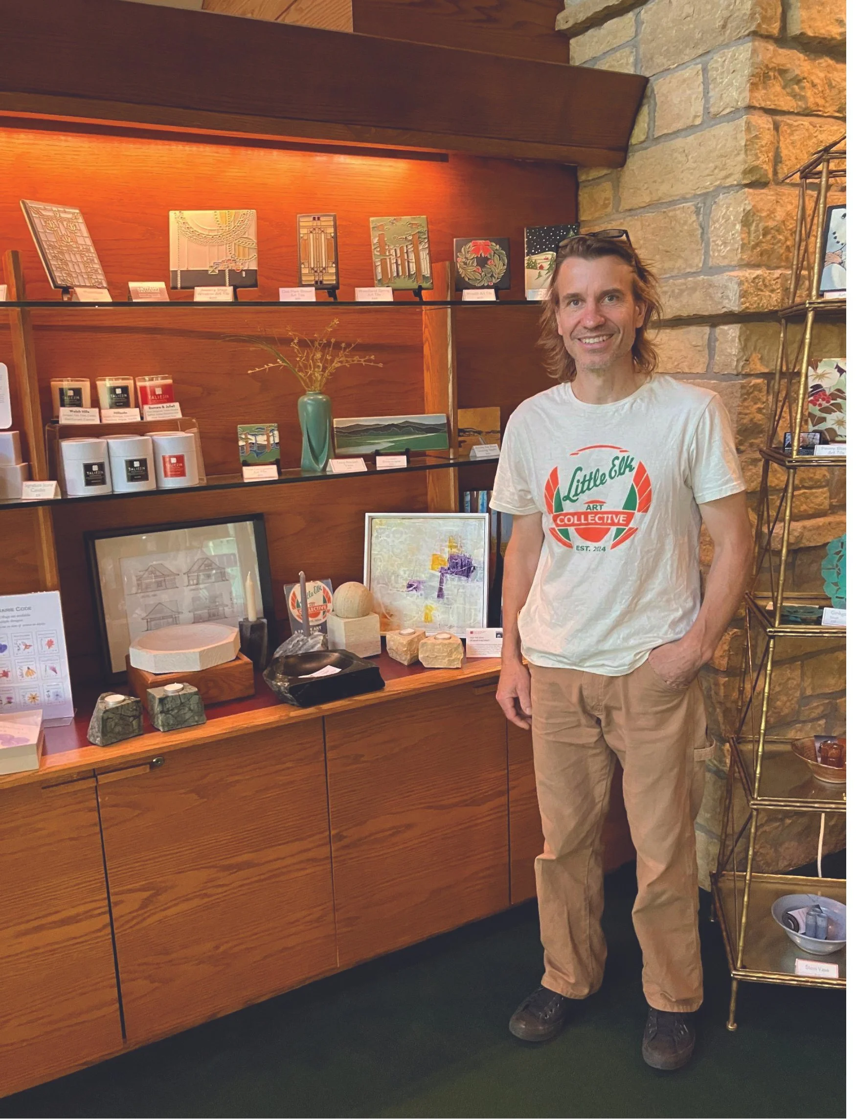 A man with long hair in a white 'Little Elk Art Collective' t-shirt and beige pants standing indoors next to wooden shelving, displaying various art pieces, ceramics, and decorative objects in a store or gallery setting.