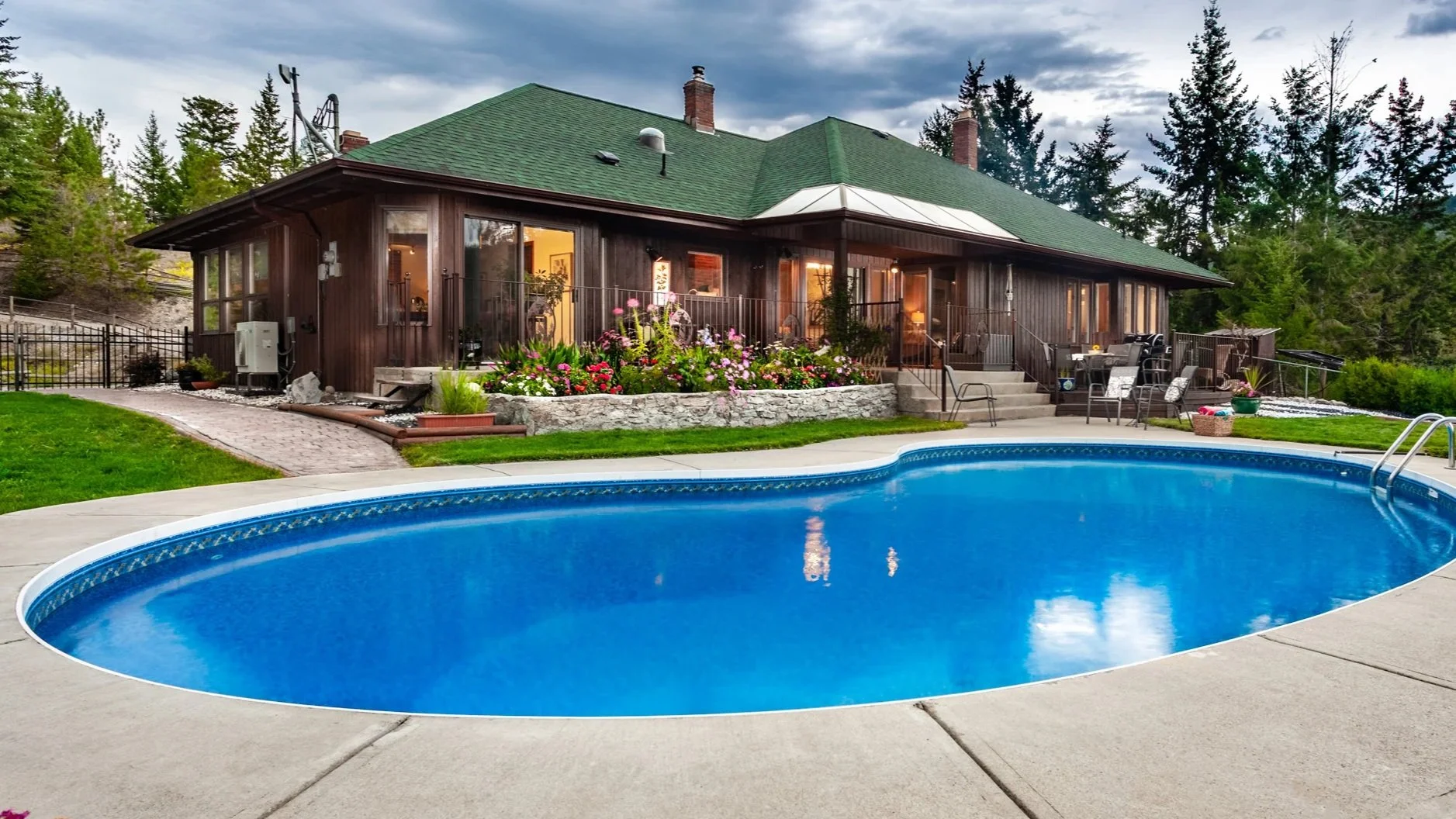 A backyard with a swimming pool, a concrete patio, a garden with flowers, and a wooden house with a green roof and large windows surrounded by trees.