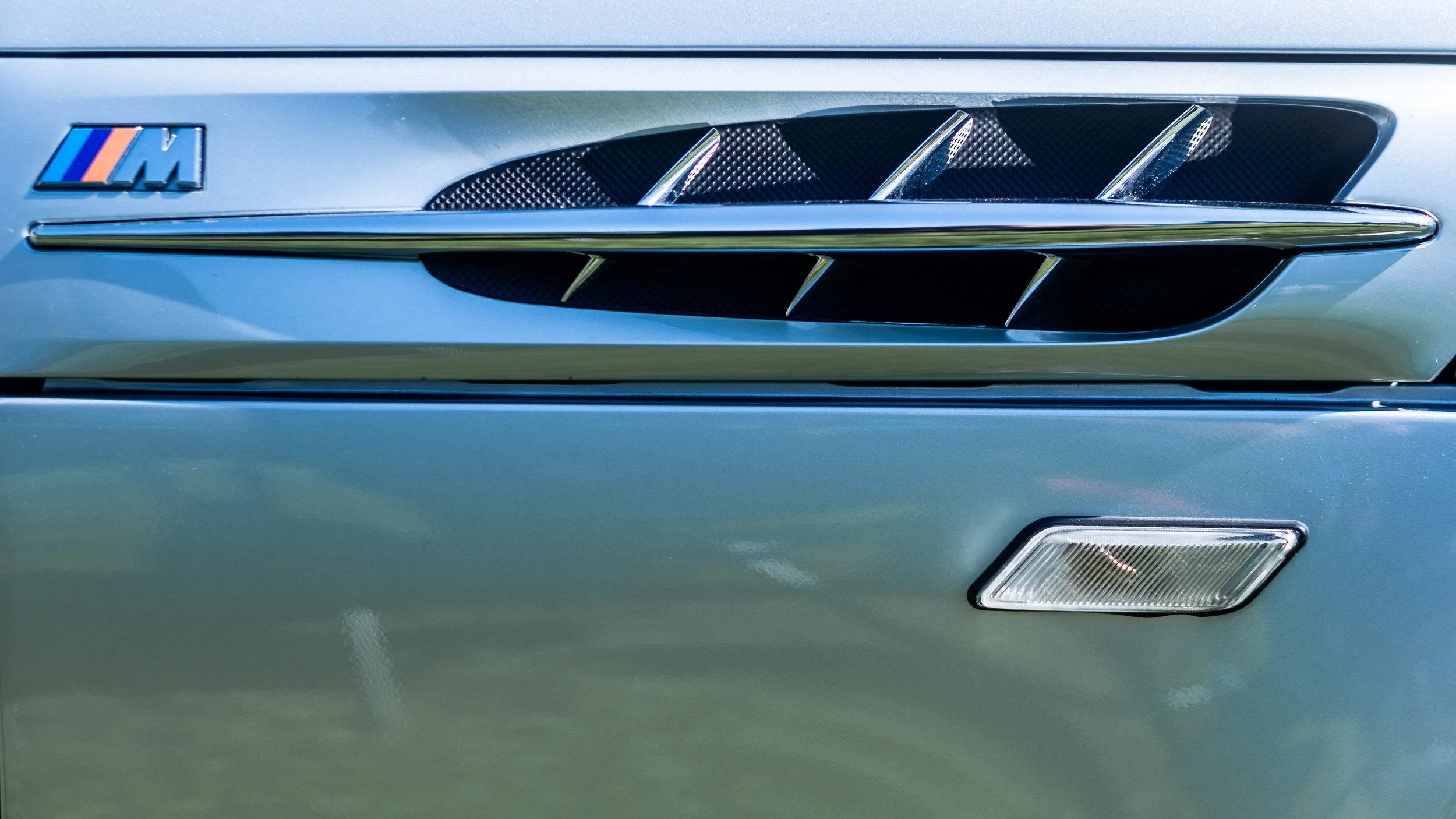 Close-up of the front side of a silver car featuring a BMW M badge and air intake vents.