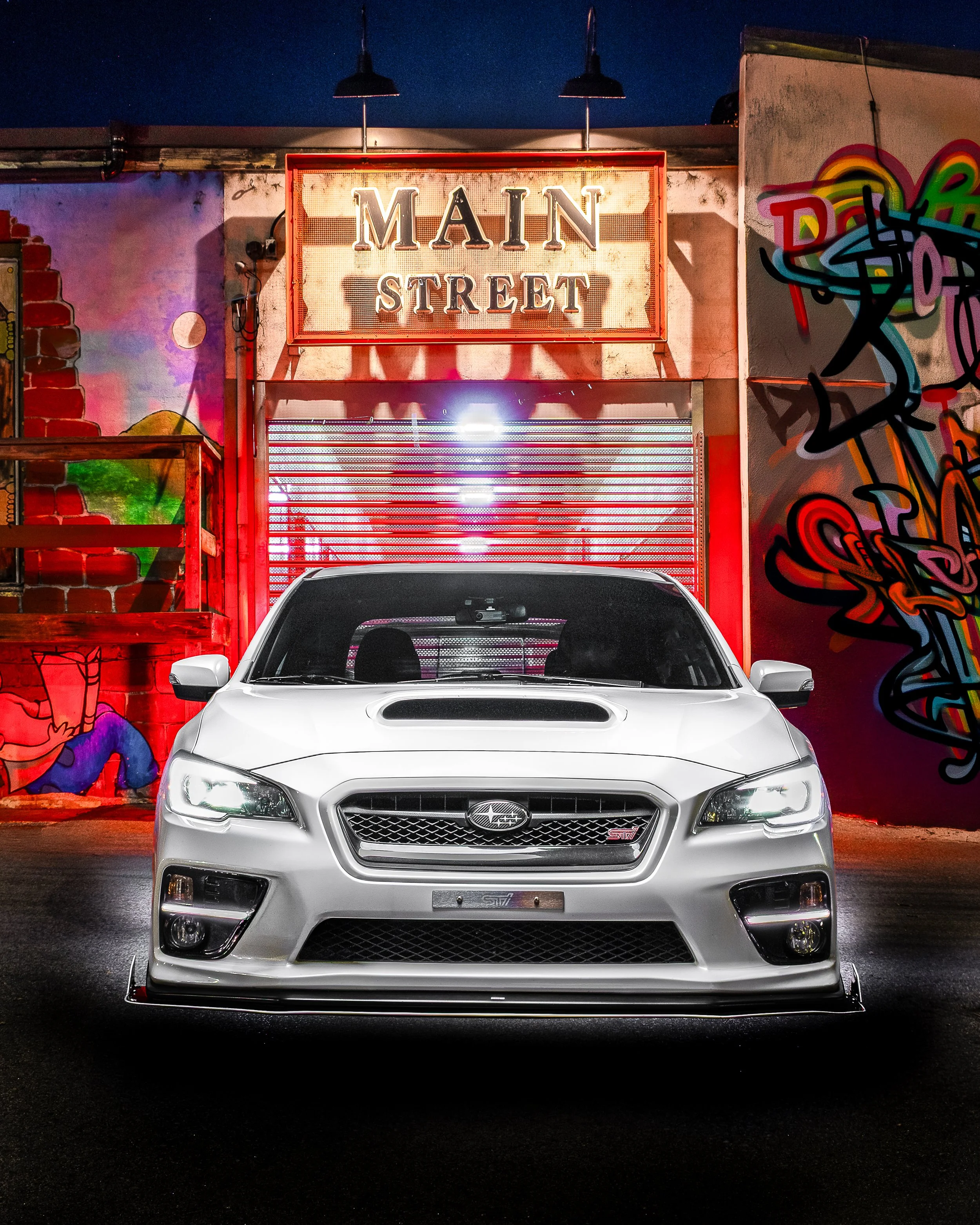 White Subaru car parked in front of a graffiti-covered wall and illuminated Main Street sign at night.