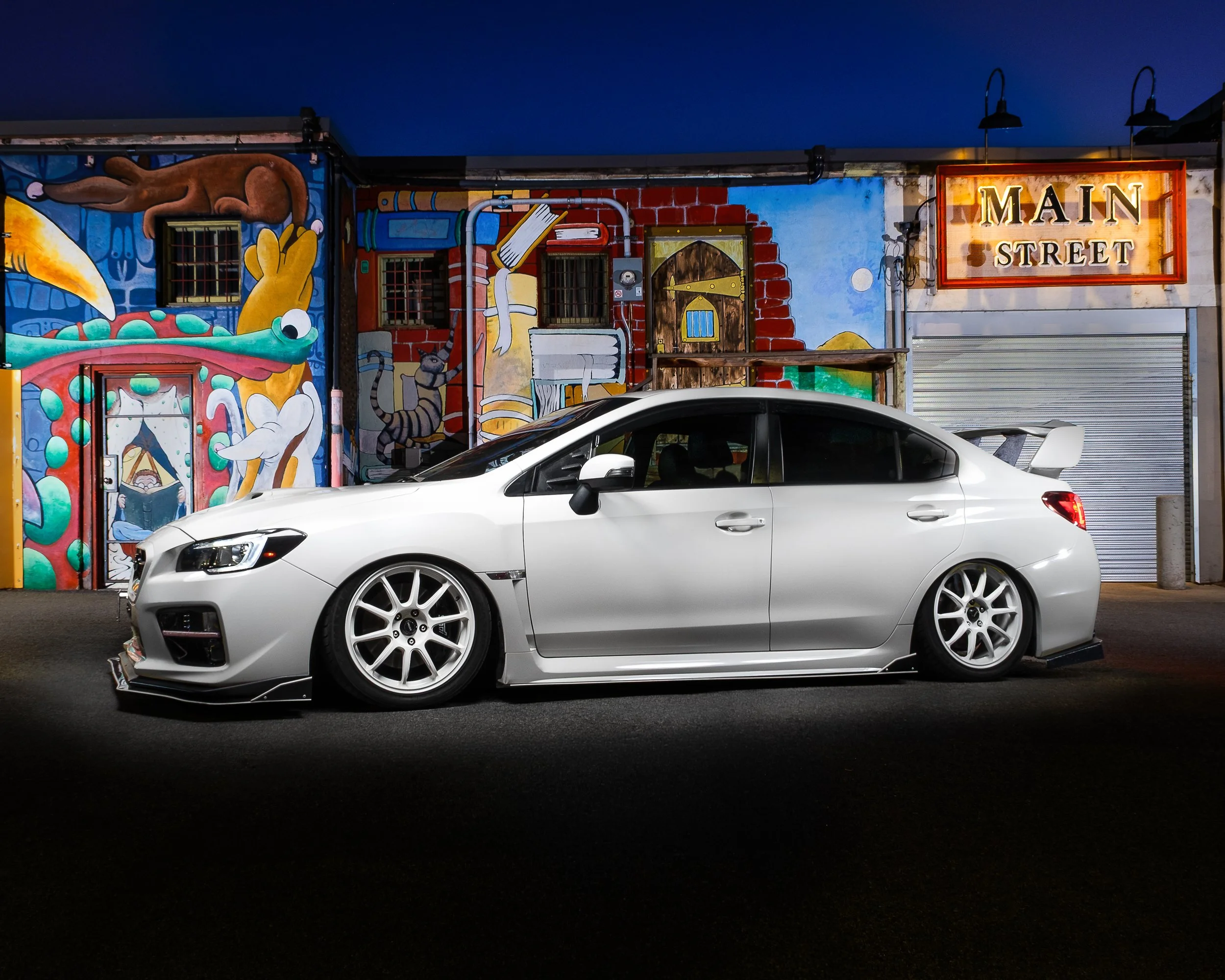 A white car parked on the street in front of a colorful mural at night, with a sign that says 'Main Street' illuminated on the building.
