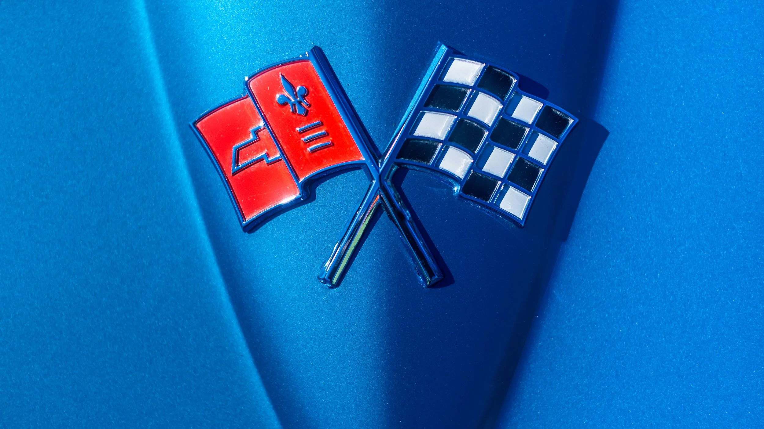 Close-up of a Cadillac emblem with a red flag featuring a blue fleur-de-lis and a blue grille design against a blue metallic surface.