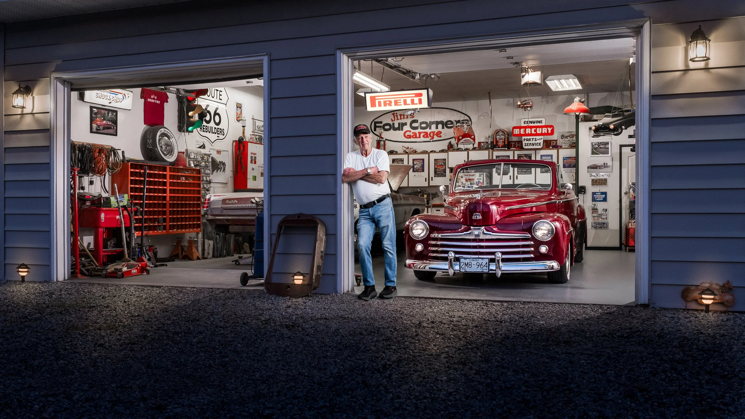 A man stands in a garage with vintage cars, automotive signs, and tools. The garage exterior shows blue siding and open garage doors.