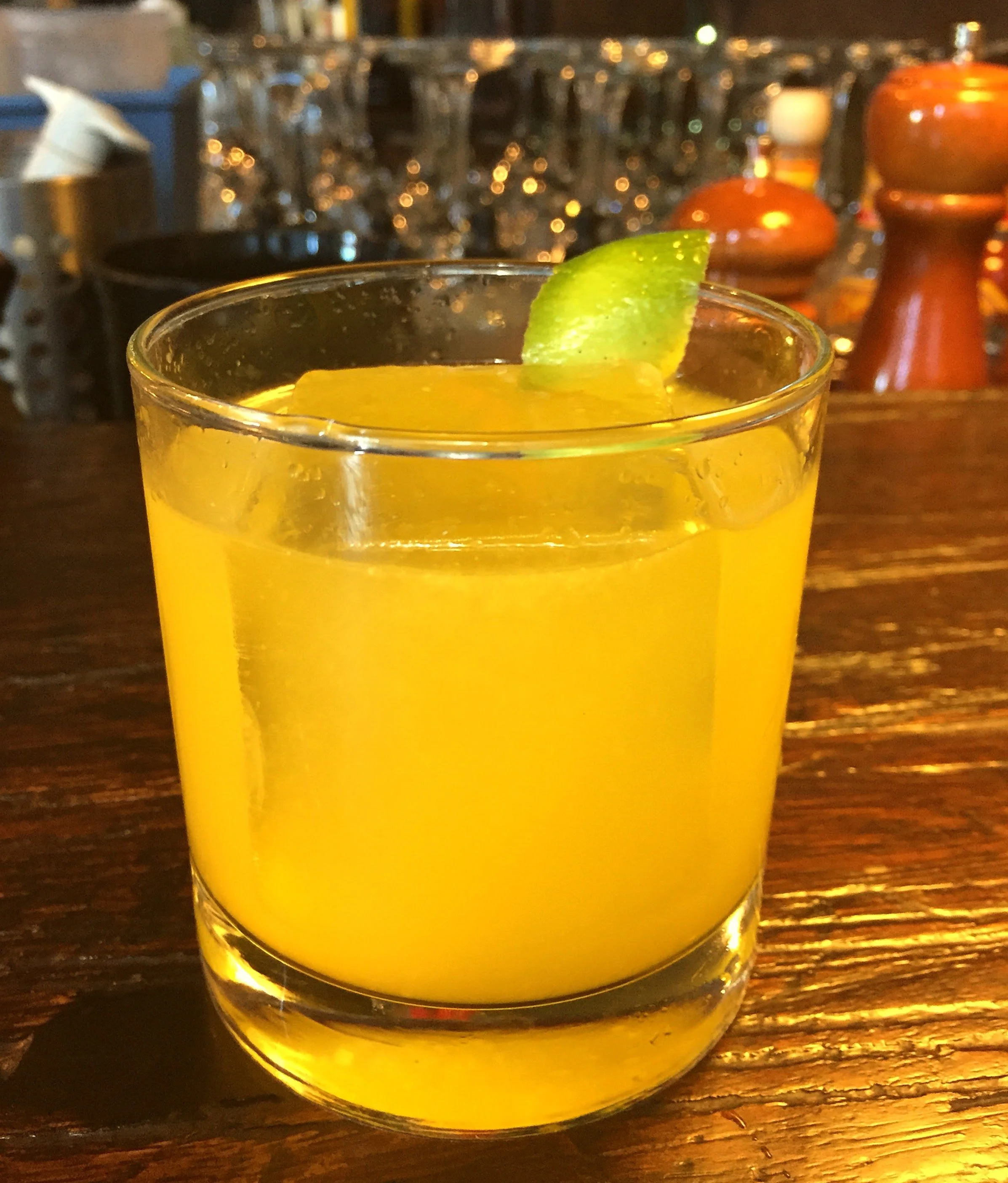 A glass of yellow-colored drink with a lime wedge on the rim, placed on a wooden table with a blurred background containing bottles and glasses.