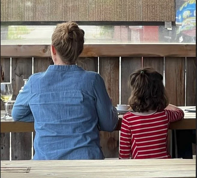 A woman and a young girl sitting at a table viewed from behind, looking out a window with a wooden fence outside. The woman is wearing a denim shirt, and the girl is wearing a red and white striped shirt.