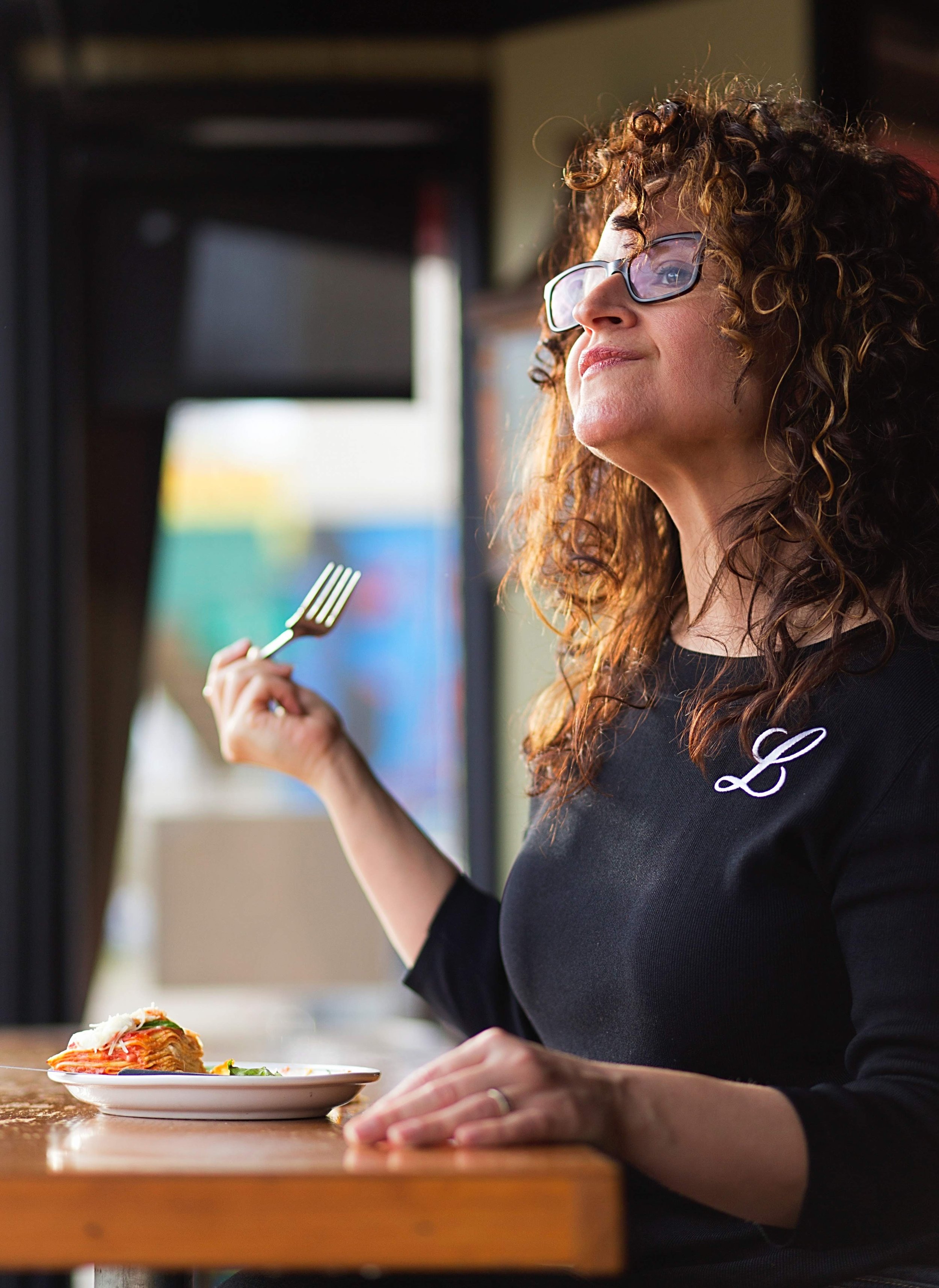 A woman with curly hair and glasses at a restaurant, holding a fork near her face, and a plate of pasta in front of her.