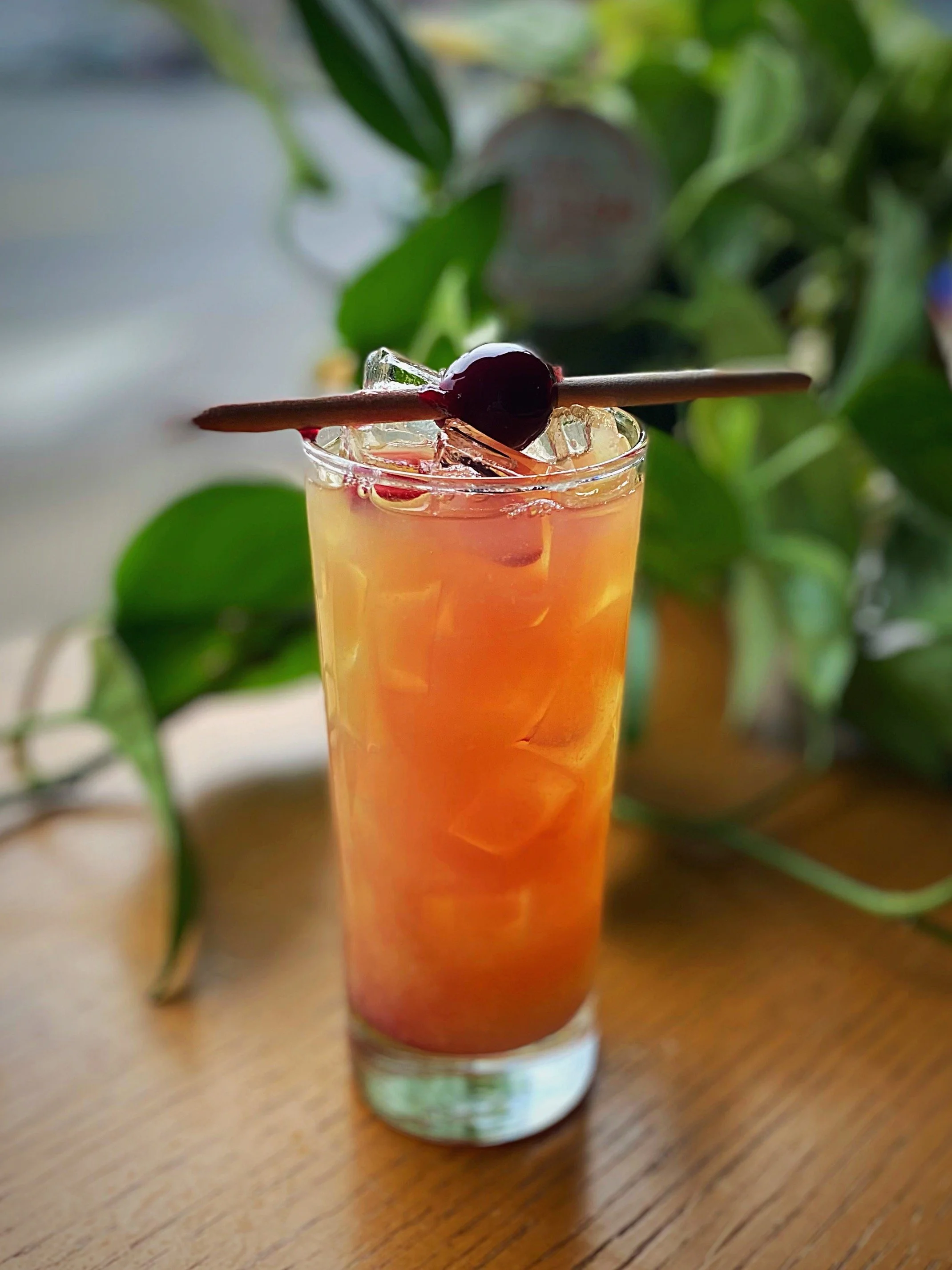 A tall glass of orange-colored beverage with ice cubes, garnished with a black cherry on a wooden stir stick, set on a wooden surface with blurred green leaves in the background.
