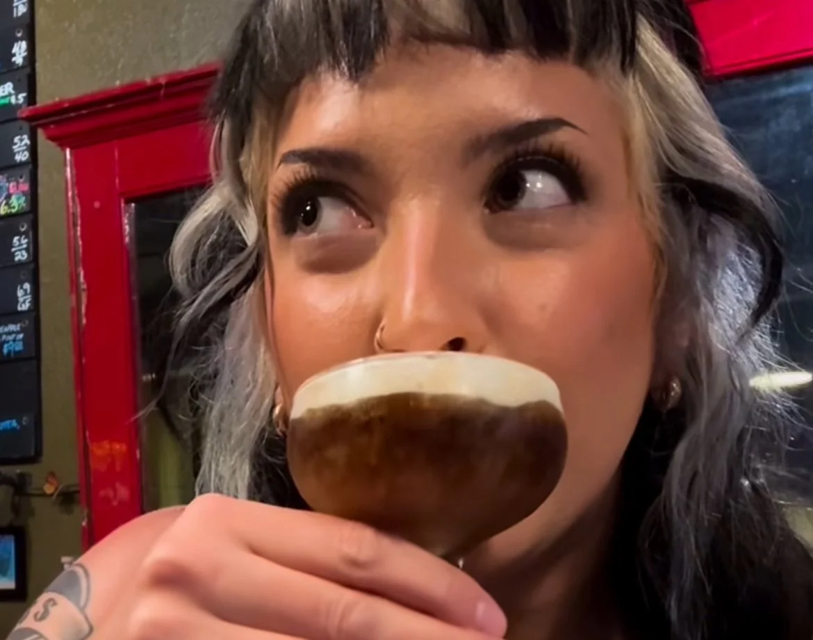A woman with gray hair and a nose piercing holds a cocktail glass filled with a dark beverage and a frothy white top, near her lips.