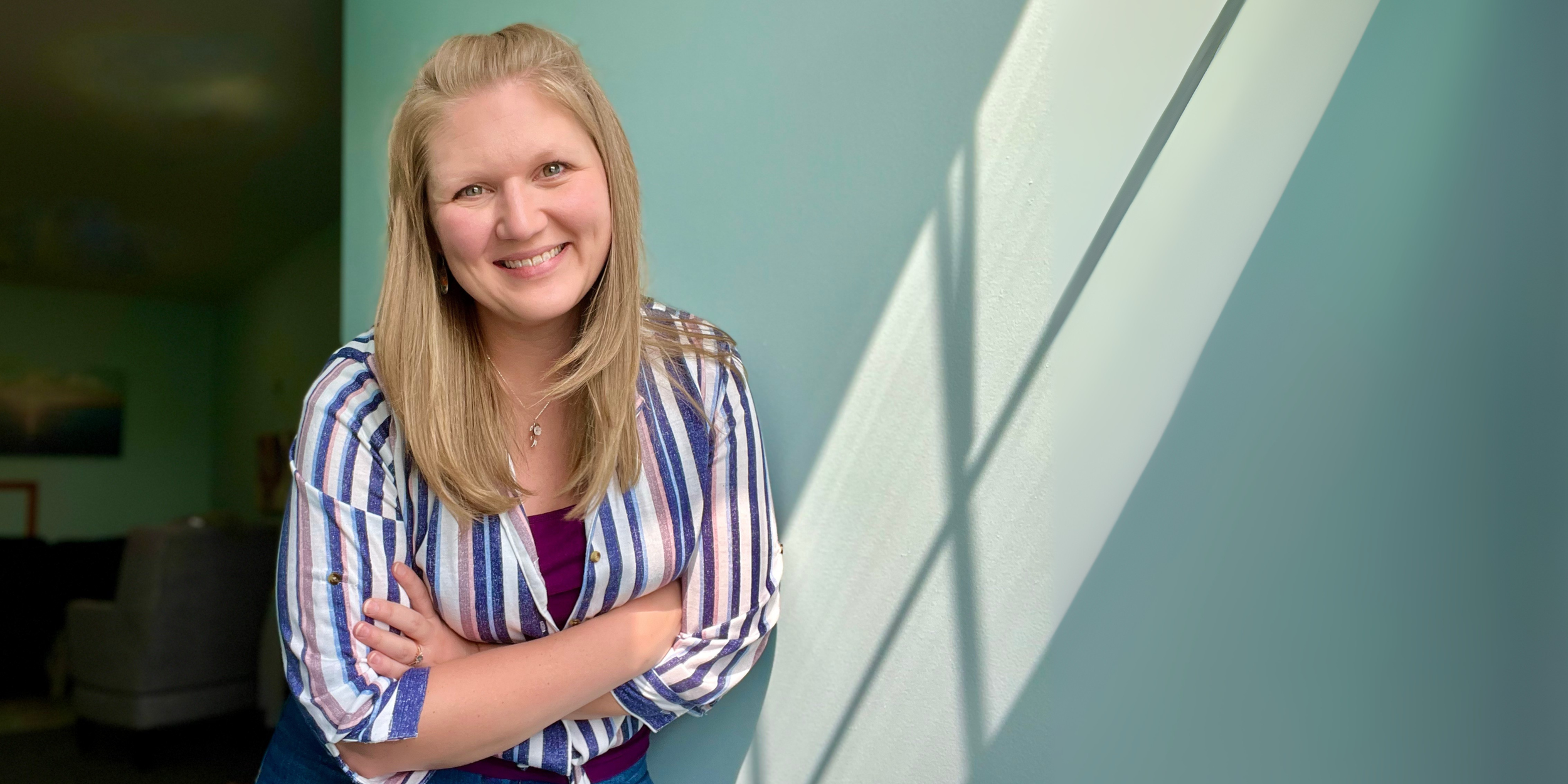 Portrait of a woman with blonde hair, smiling, wearing a striped shirt, standing against a light green wall with shadows from window blinds.
