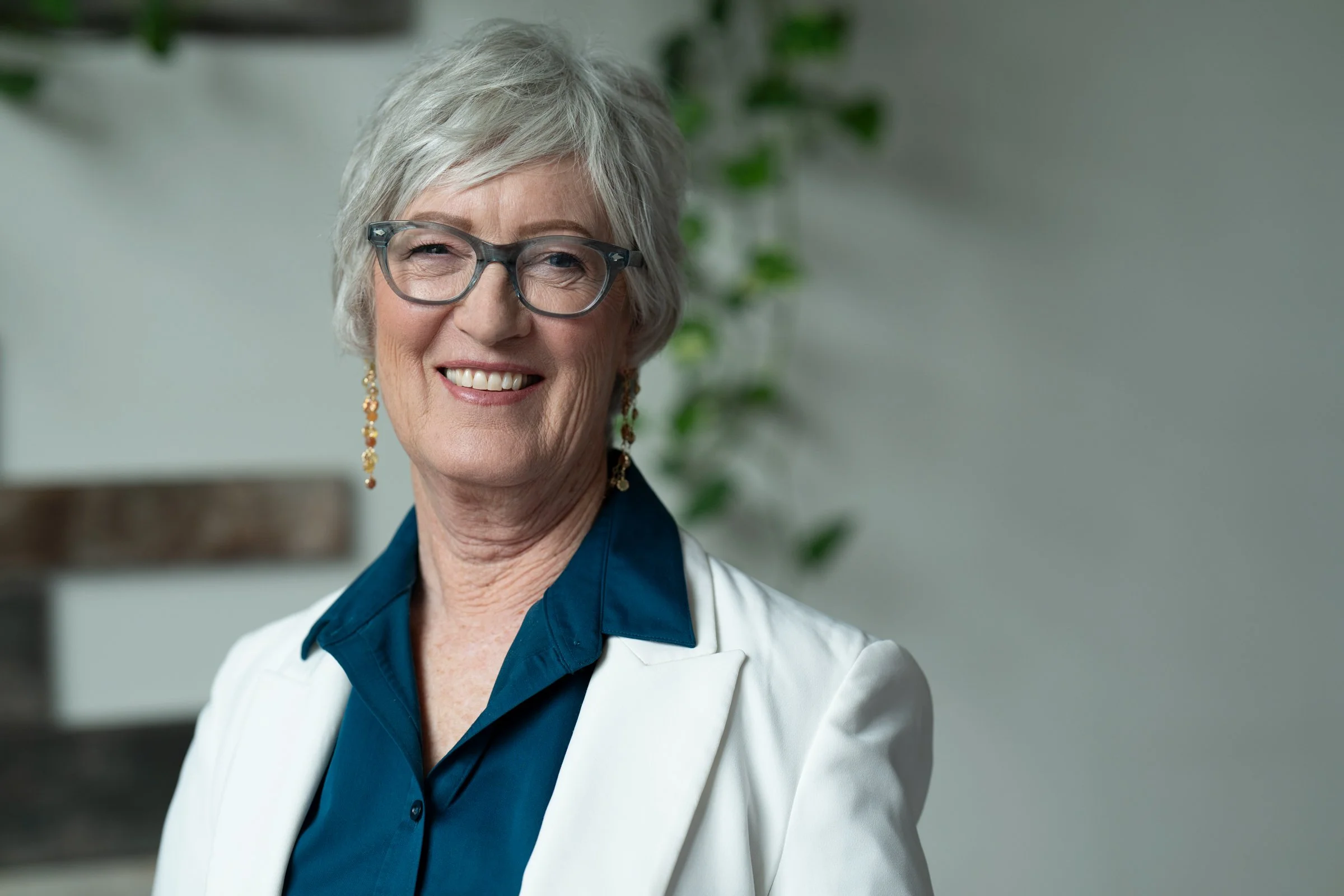 Professional executive portrait of a confident woman with short gray hair and glasses, wearing a white blazer and teal blouse, photographed in natural light.