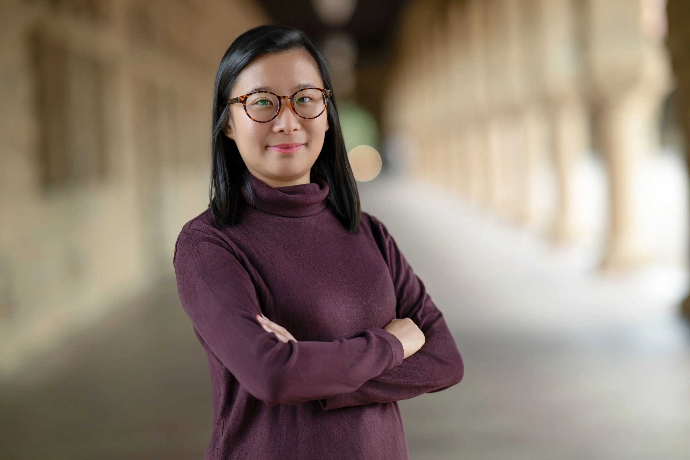 Professional portrait of a woman wearing glasses and a burgundy top, standing with arms crossed in a softly blurred architectural hall