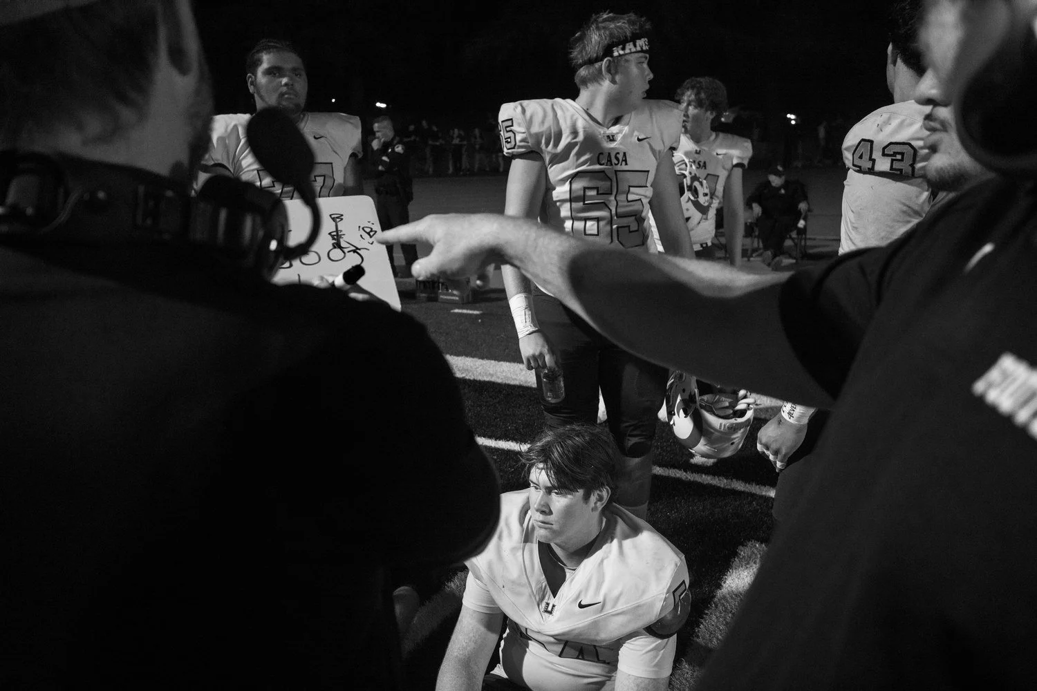 Black-and-white sideline shot of a coach pointing past a camera operator toward a seated football player at night