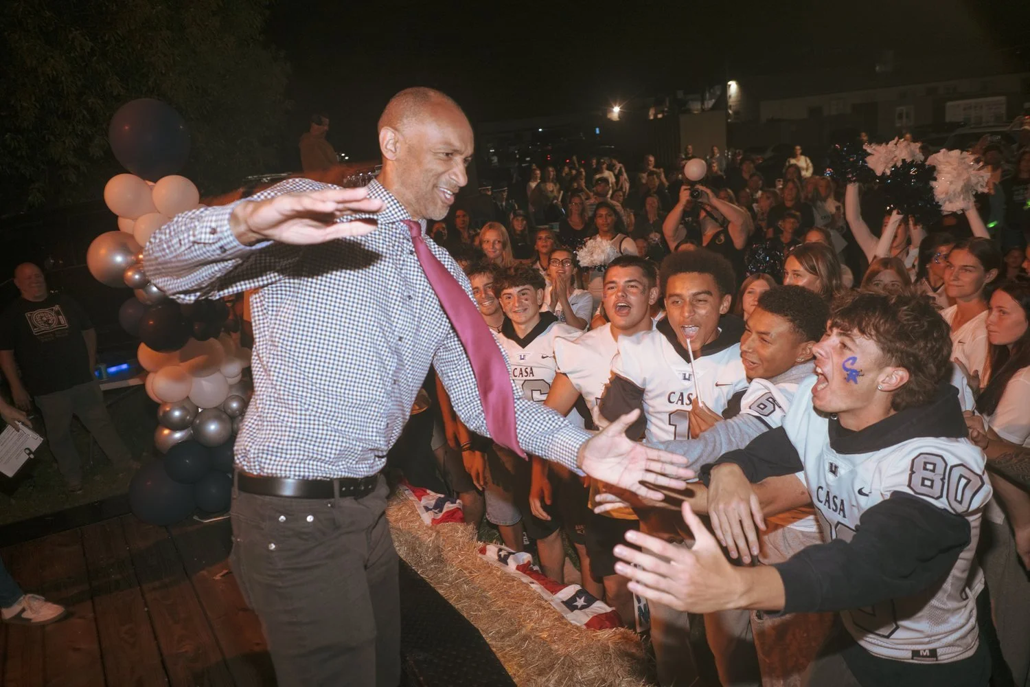 A man in a shirt and tie high-fives a packed, cheering student section at a night football game