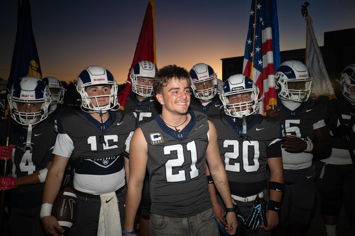 Smiling football players in helmets stand in front of flags under an orange dusk sky before a game