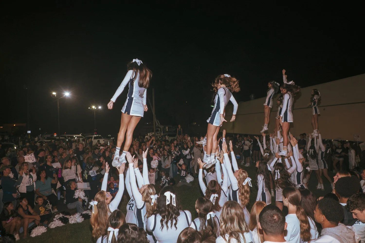 Cheerleaders held up in stunts above a large crowd under stadium lights at a night football game