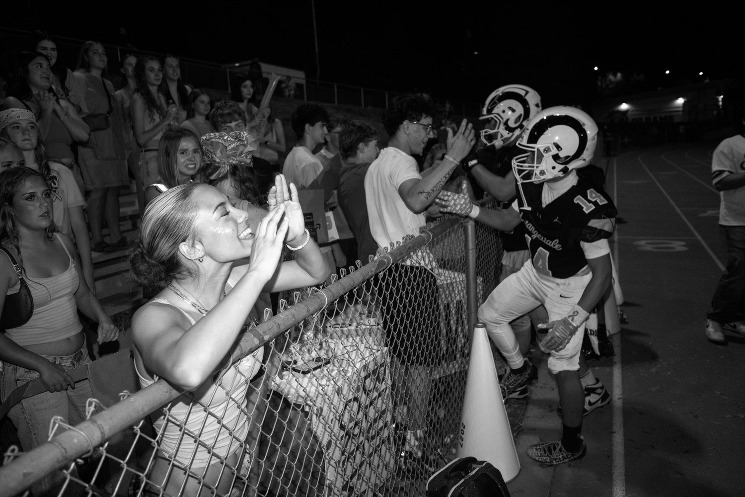 Black-and-white photo of fans at a chain-link fence reaching toward football players walking past at night
