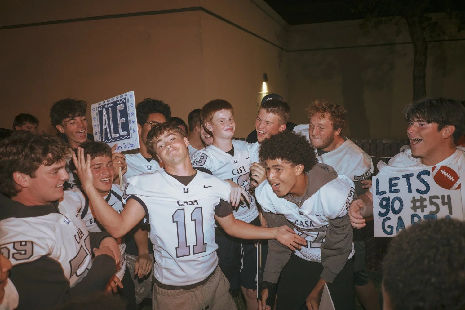 Excited high school students in jerseys crowd together holding handmade signs, lit by on-camera flash