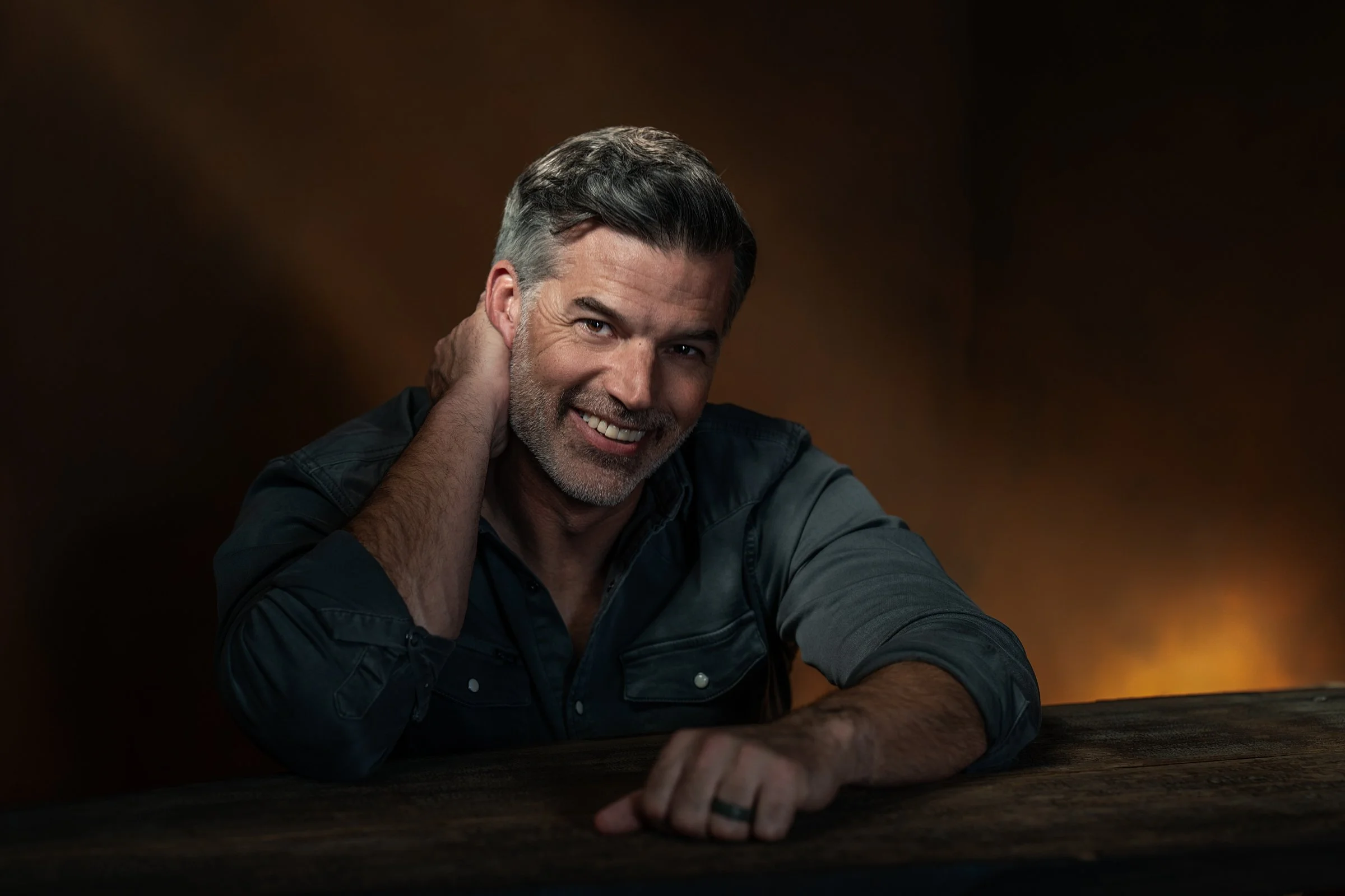 Professional portrait of a smiling man with salt-and-pepper hair leaning forward, photographed in a studio with warm, dramatic lighting.